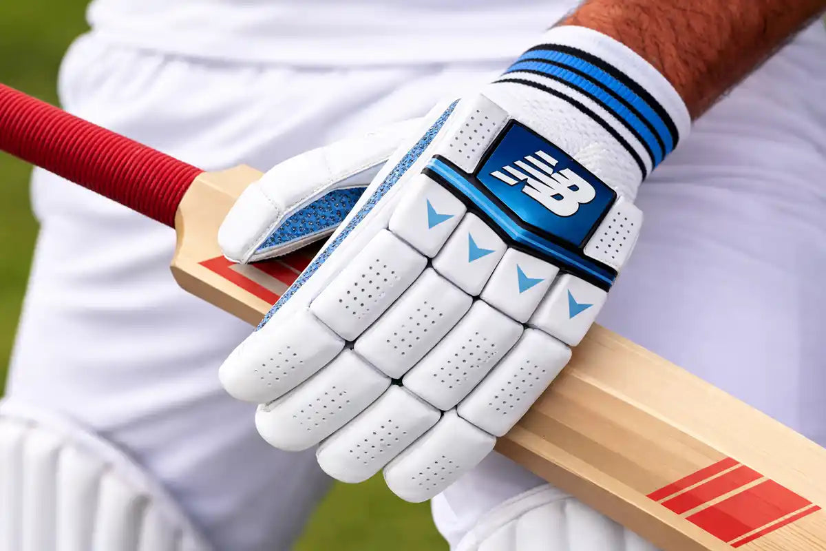 Close-up of a cricket player wearing New Balance batting gloves gripping a wooden bat, highlighting enhanced grip, control, and protective padding during play